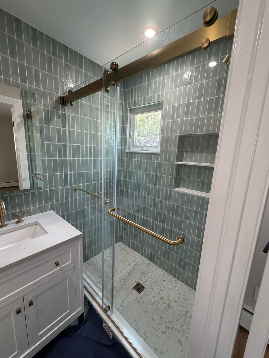 Shower after renovation: green stacked tile, recessed niche under the window, brass sliding glass door and terrazzo-style floor