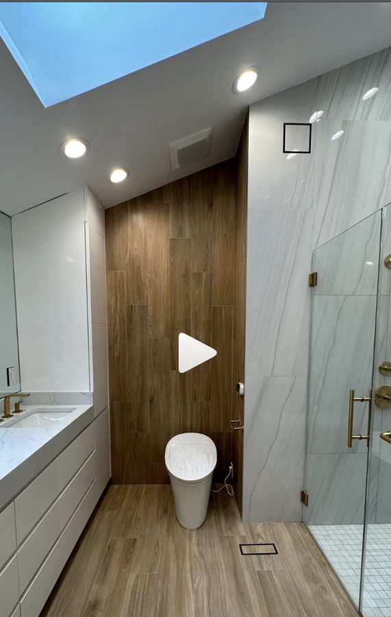 Primary bathroom with wood-look tile accent wall, frameless glass shower, and brass fixtures